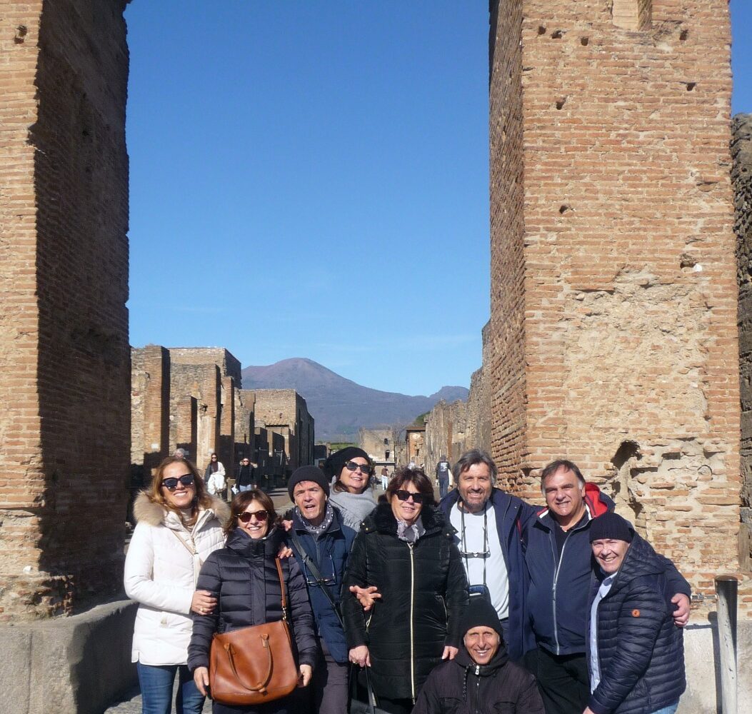 A group of tourists posing under a large ancient Roman brick archway in Pompeii with Mount Vesuvius in the background.