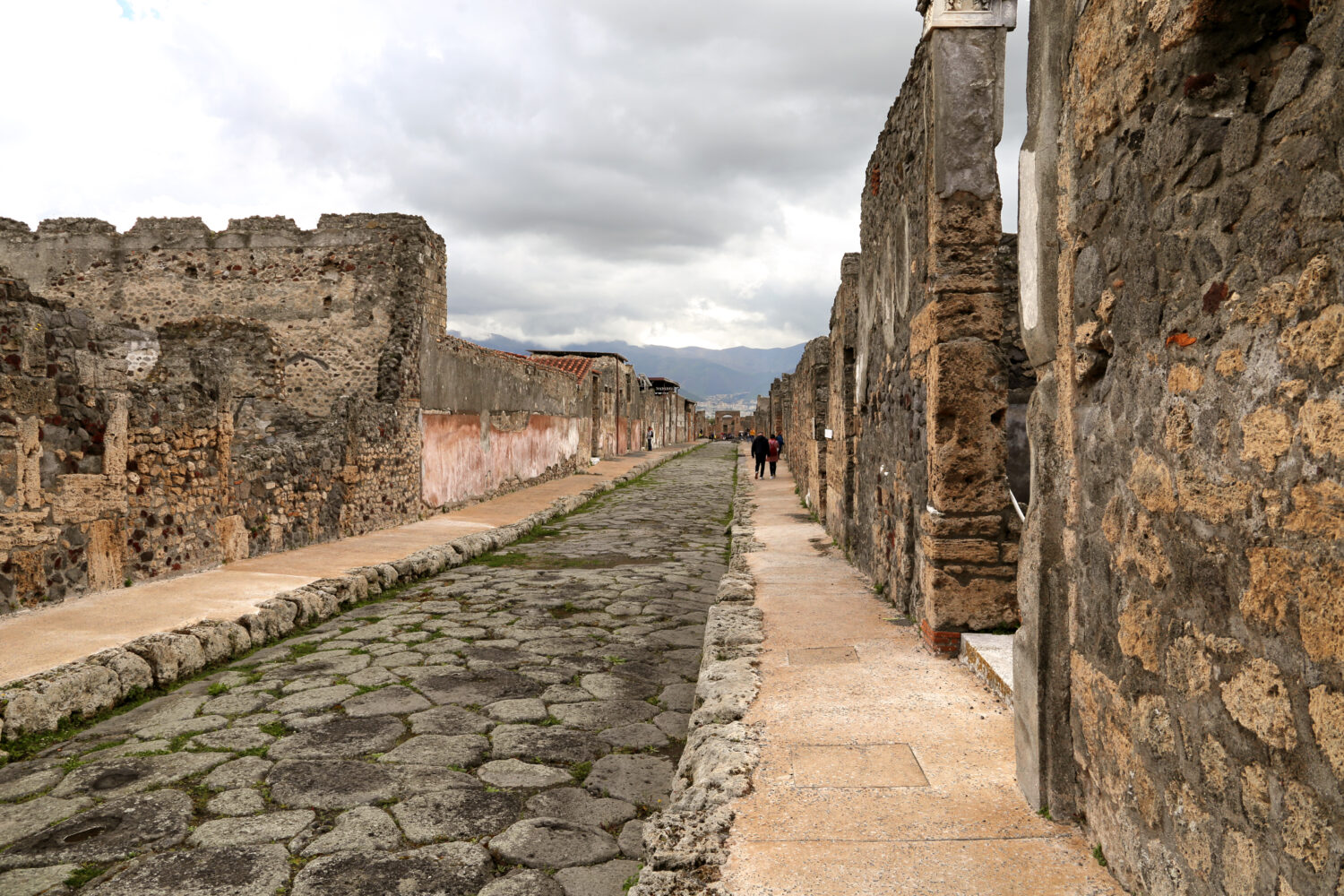 Long perspective of an ancient Roman street in Pompeii with stone walls and distant mountains.
