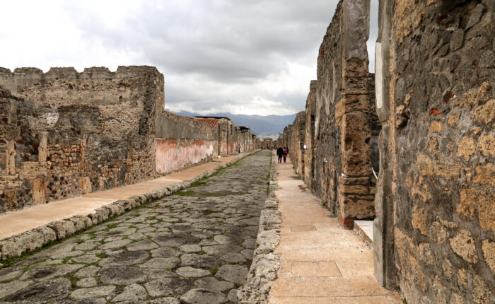 Long perspective of an ancient Roman street in Pompeii with stone walls and distant mountains.