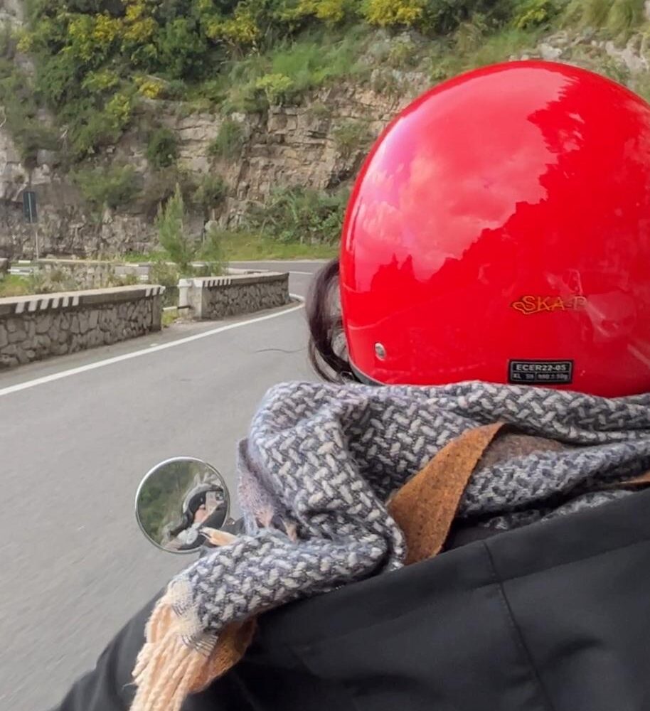 Person with a red helmet riding a scooter on a scenic coastal road along rocky cliffs.